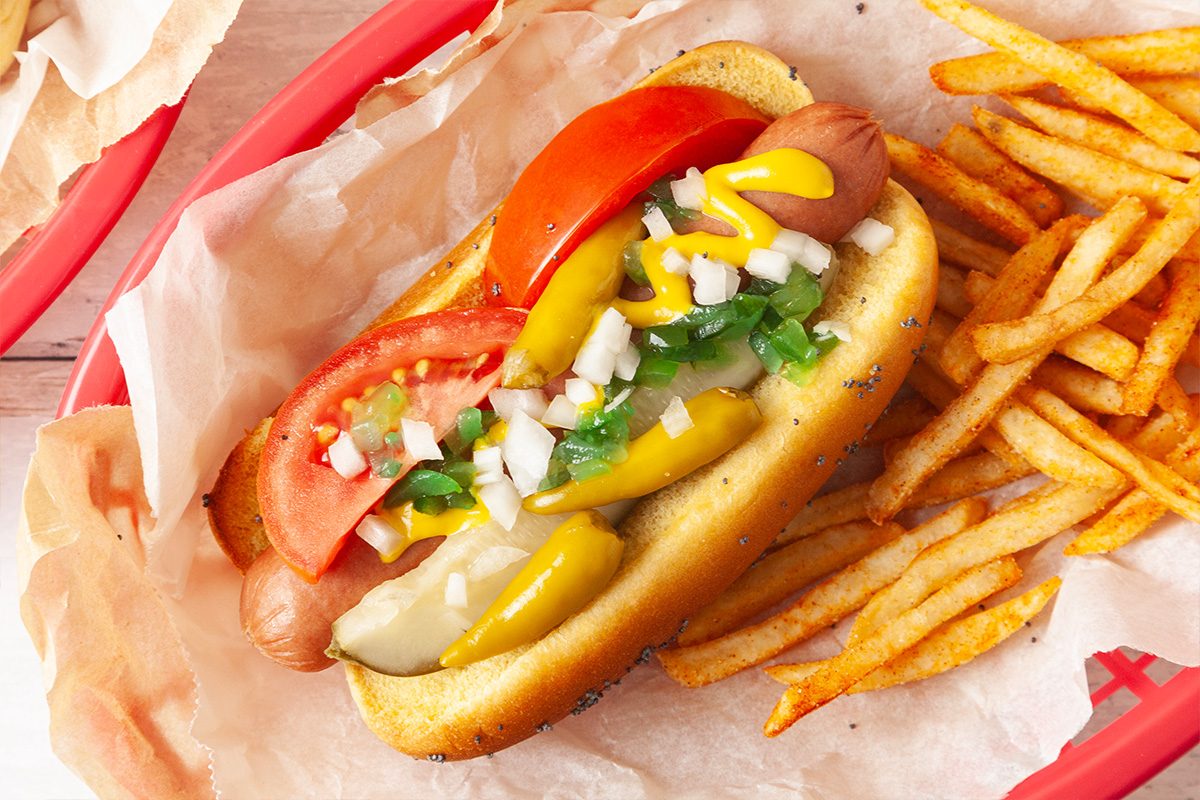 A Chicago-style hot dog topped with tomato slices, pickles, onions, peppers, relish, and mustard in a bun, served with a side of seasoned French fries in a red basket.