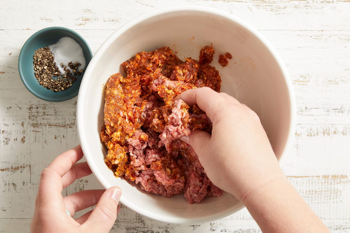 A person mixes ground meat with spices in a white bowl. A small blue bowl containing salt and pepper sits nearby on a white wooden surface.