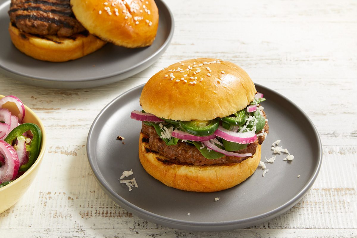 A grilled burger patty on a sesame bun topped with sliced red onions, green peppers, and lettuce sits on a gray plate. Another burger and a bowl of sliced vegetables are in the background.