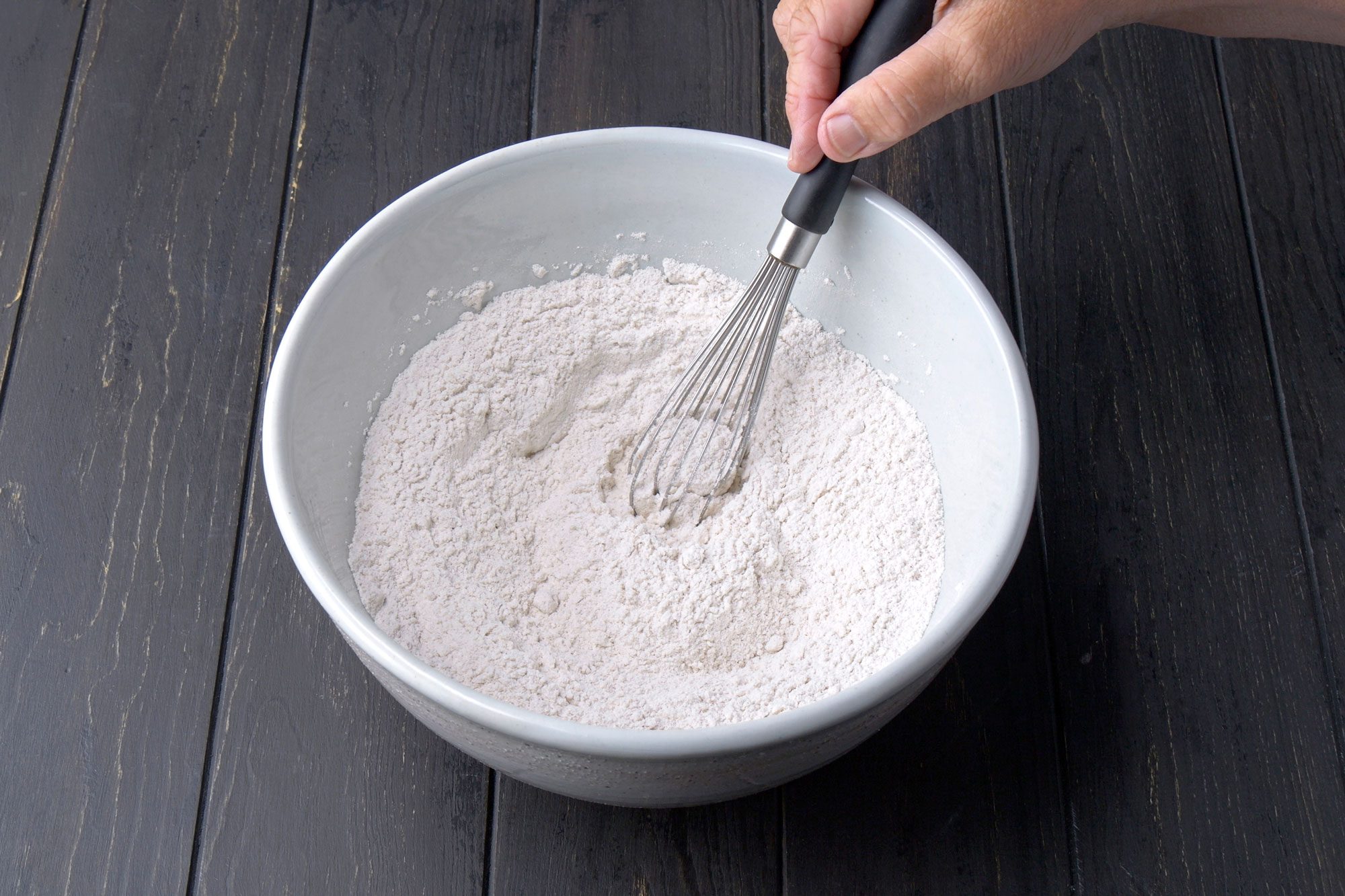3/4th shot of a hand whisking flour in a large white bowl on a dark wooden surface