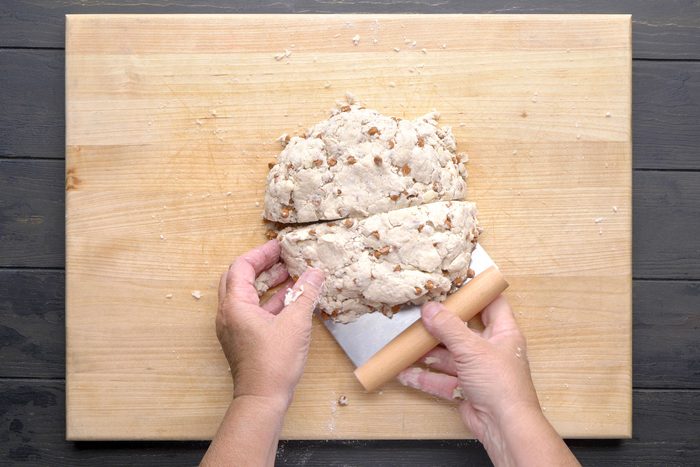 overhead shot of A person uses a bench scraper to cut a round ball of dough with nuts on a wooden cutting board; The board sits on a dark countertop