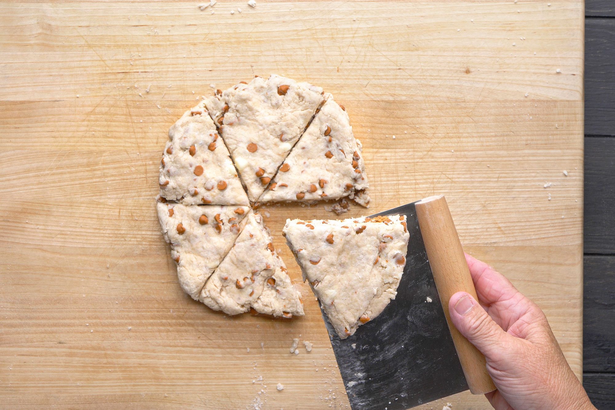 overhead shot of hand uses a dough scraper to lift a wedge shaped piece of unbaked dough with chocolate chips from a round, divided dough on a wooden cutting board