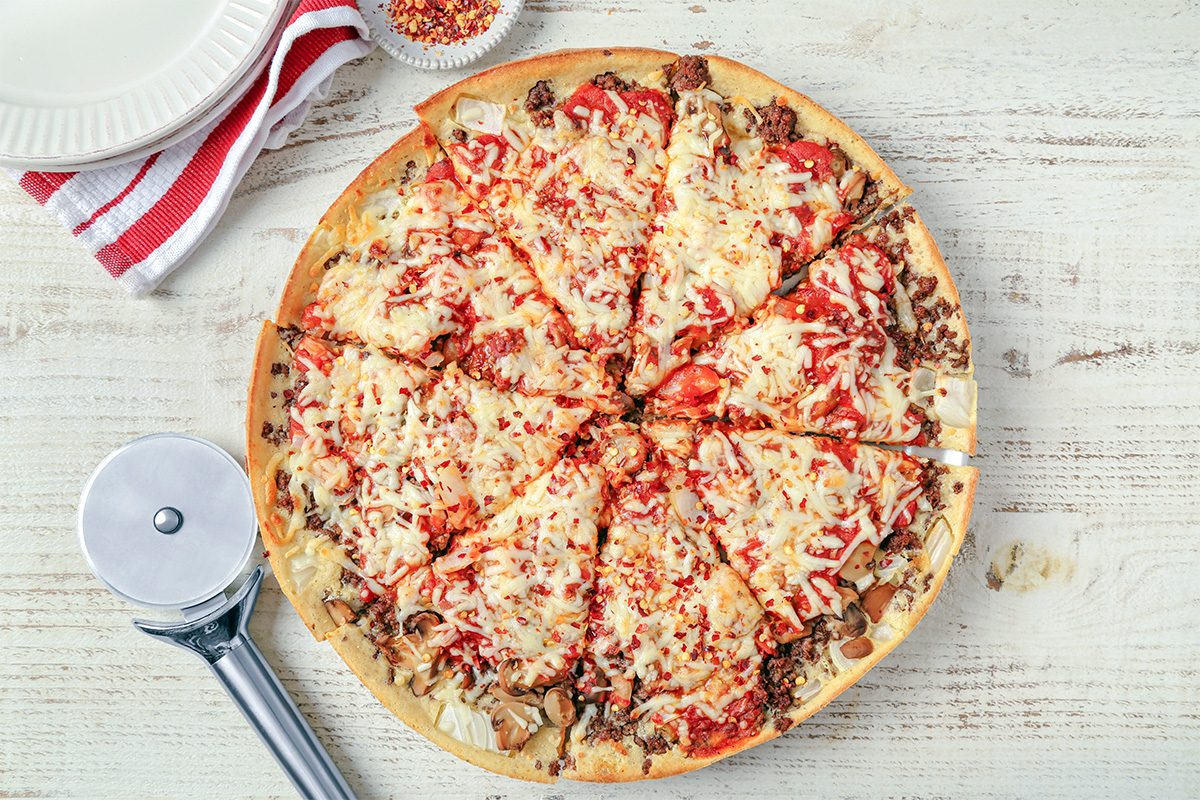 A whole pizza topped with cheese, ground meat, and tomato sauce is sliced into eight pieces on a wooden surface. A pizza cutter, a striped napkin, plates, and a bowl of crushed red pepper are nearby.