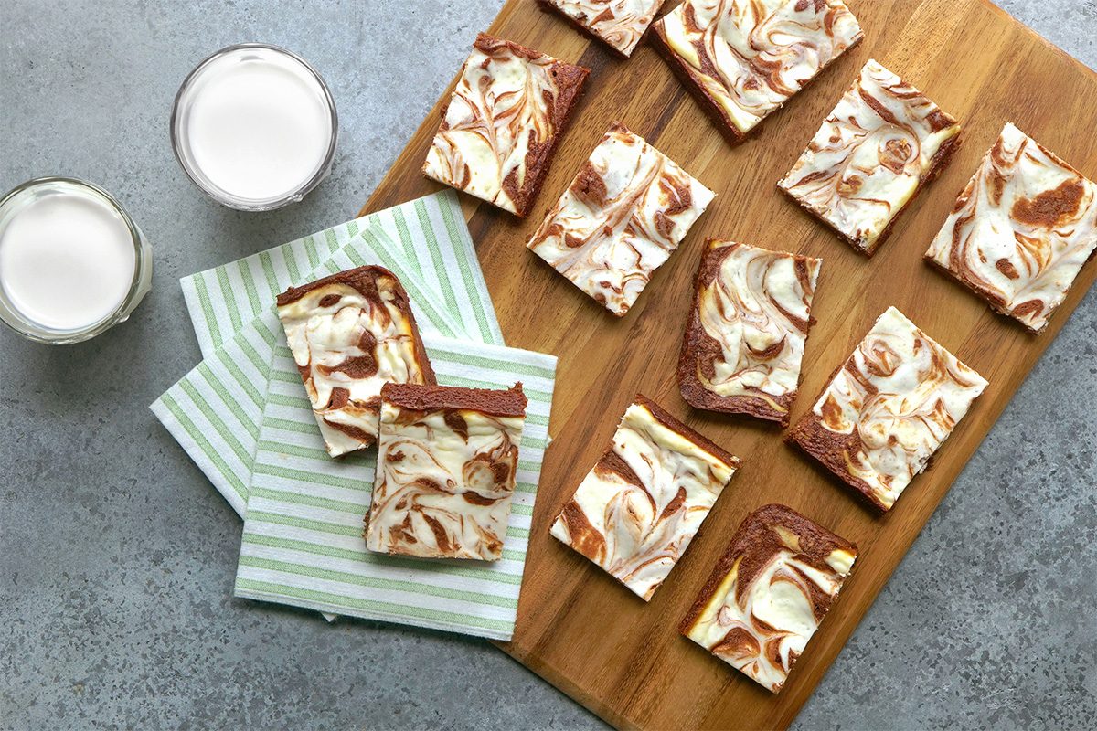 A wooden board with several pieces of marbled cheesecake brownies, two placed on striped napkins next to two glasses of milk, all on a gray surface.