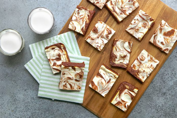 A wooden board with several pieces of marbled cheesecake brownies, two placed on striped napkins next to two glasses of milk, all on a gray surface.