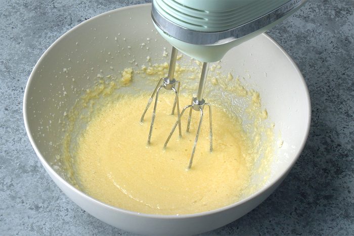 A hand mixer blends a pale yellow batter in a white mixing bowl on a gray countertop.