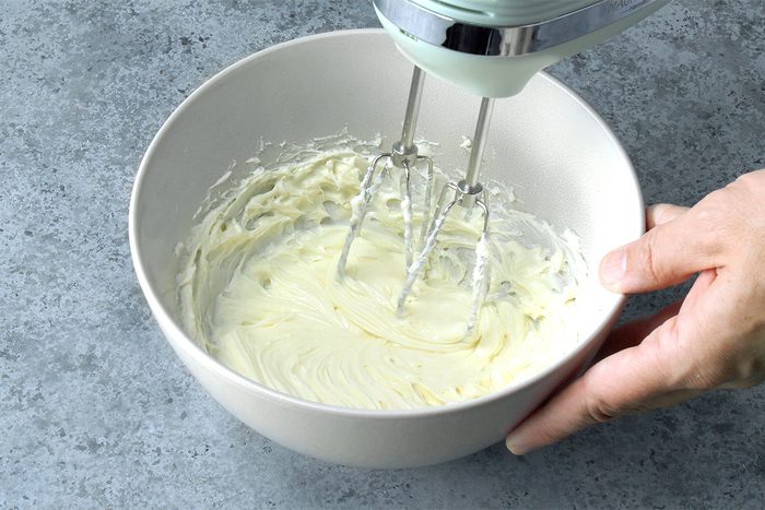A hand holds a white mixing bowl while an electric hand mixer blends creamy batter on a gray countertop.