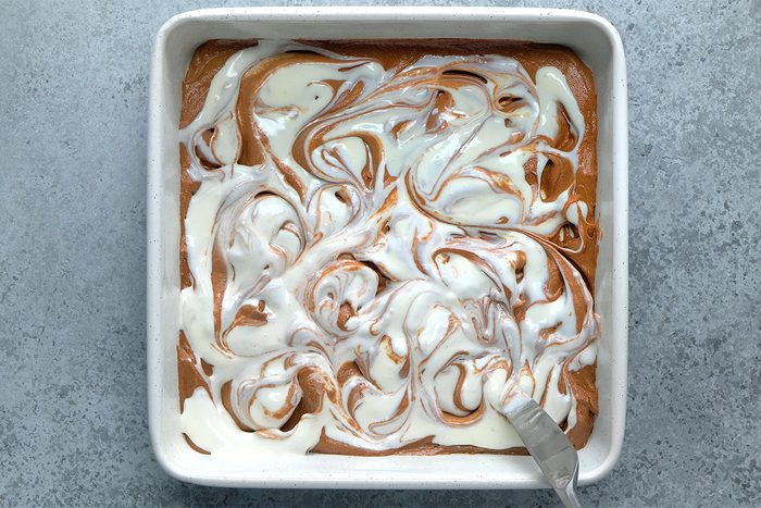 A square baking dish filled with swirled chocolate and white batter, with a spatula resting in one corner, on a gray surface.