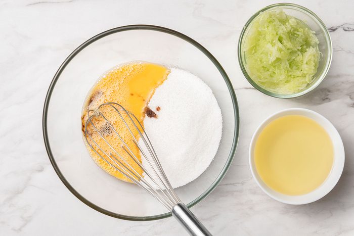 Sugar, oil, vanilla extract and grated, seeded and peeled cucumbers with eggs in bowls.