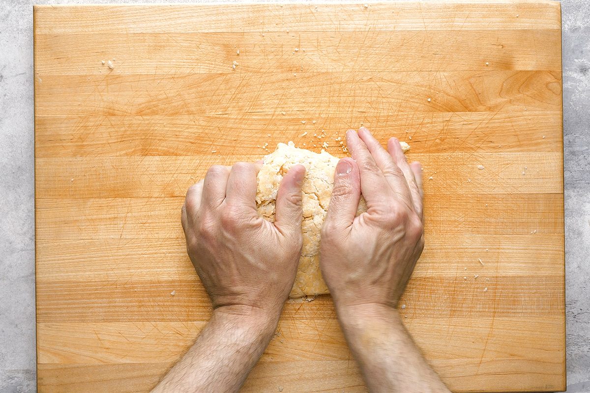 Two hands knead a mound of dough on a wooden cutting board, viewed from above. The dough appears rough and crumbly, and the board has visible grain patterns and a few scattered crumbs.