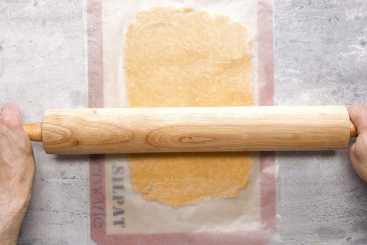 A person rolls out dough between two sheets of parchment paper using a wooden rolling pin on a lightly floured surface.