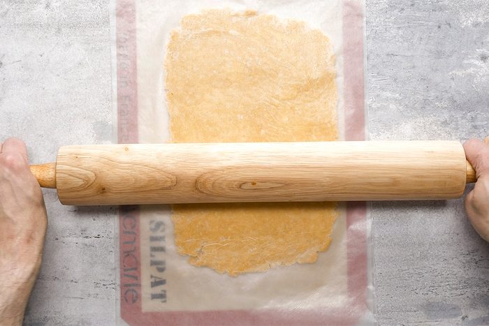 A person rolls out dough between two sheets of parchment paper using a wooden rolling pin on a lightly floured surface.