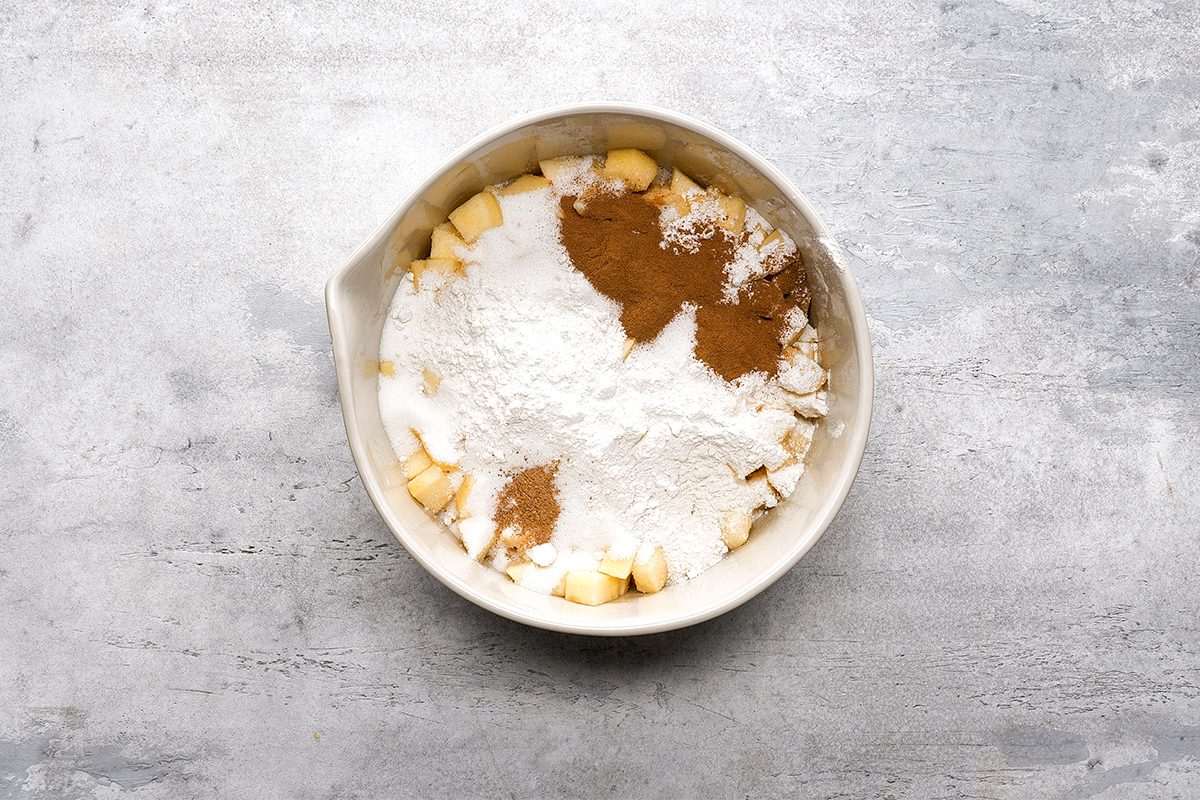 A mixing bowl filled with chopped apples, flour, sugar, and cinnamon, ready to be mixed, on a light gray textured surface.