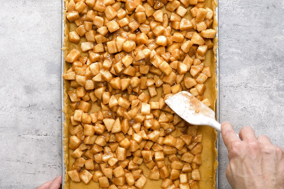 A hand spreads a layer of diced, cinnamon-coated apples onto a rectangular sheet of dough in a baking pan.