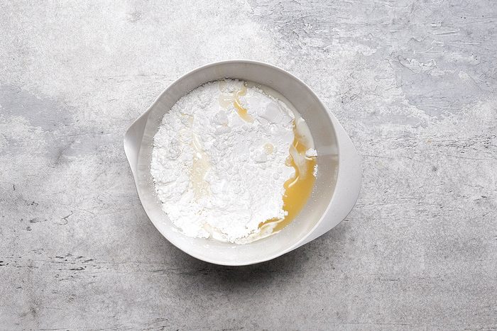 A white mixing bowl contains flour and a pool of yellow liquid, likely oil or melted butter, on a gray textured surface.