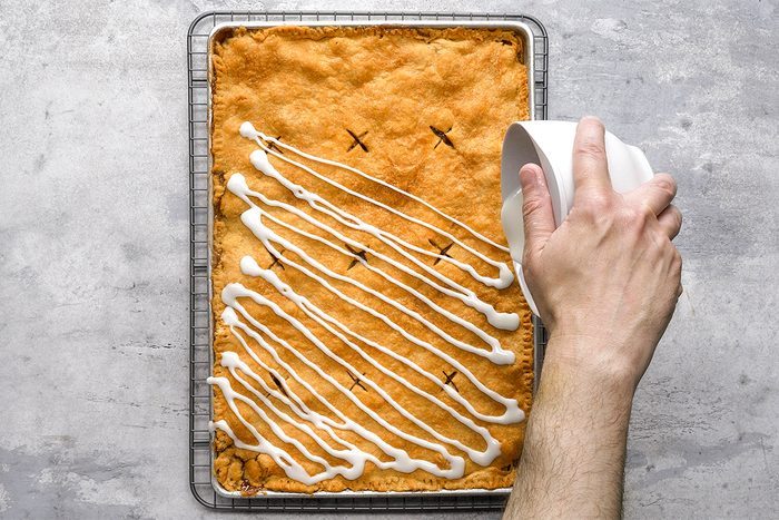 A hand drizzles white icing in diagonal lines over a large rectangular baked pastry on a cooling rack, with a few cuts visible on the golden-brown surface.