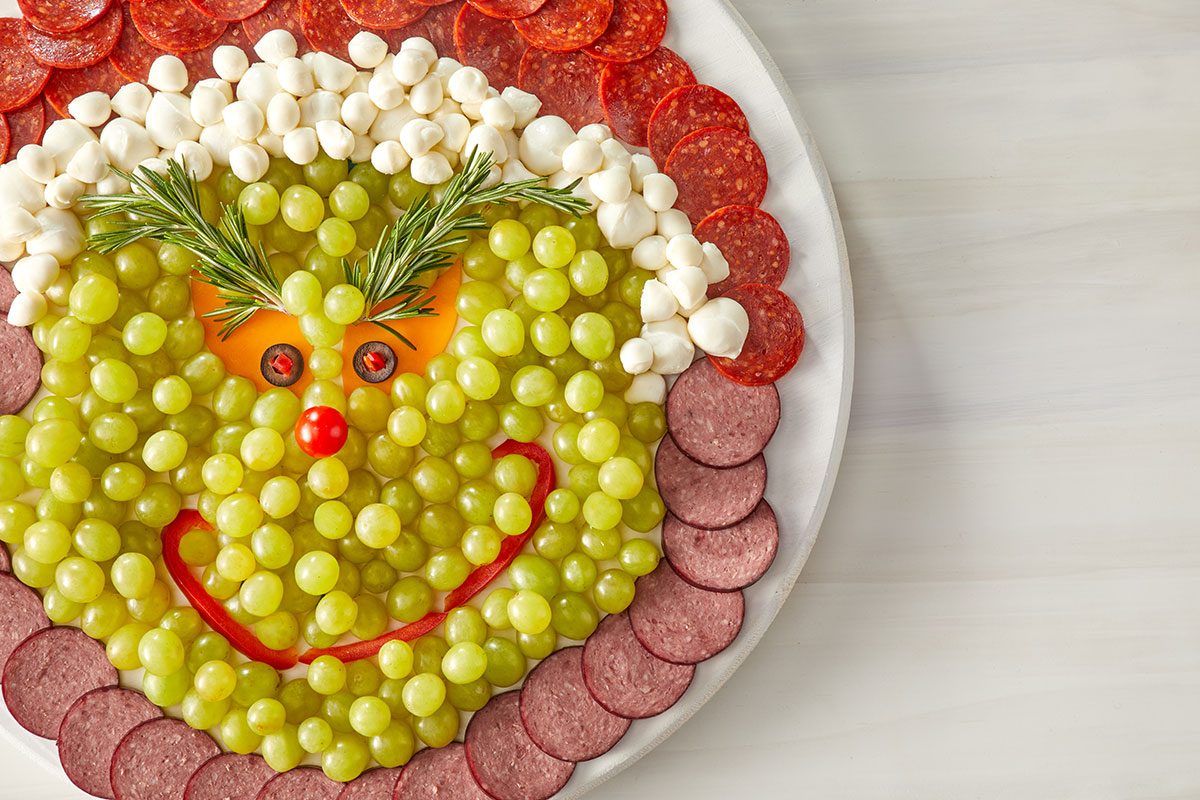Overhead shot of a festive platter arranged to look like a smiling grinch face with green grapes, cheese, meat slices, and rosemary for eyebrows on a white plate, The face has a red pepper smile and a cherry tomato nose