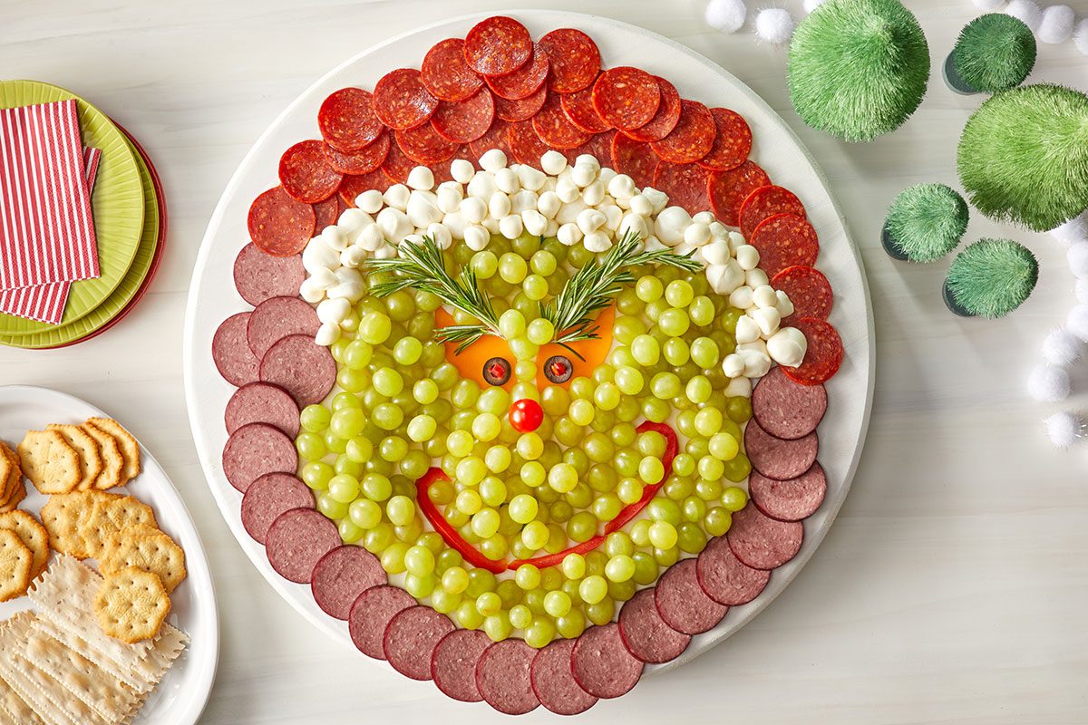 Overhead shot of a festive platter arranged to resemble the Grinch’s face, made with green grapes, cheese balls, meats, red bell pepper, and rosemary sprigs for eyebrows, Plates of crackers and napkins are nearby on a white table