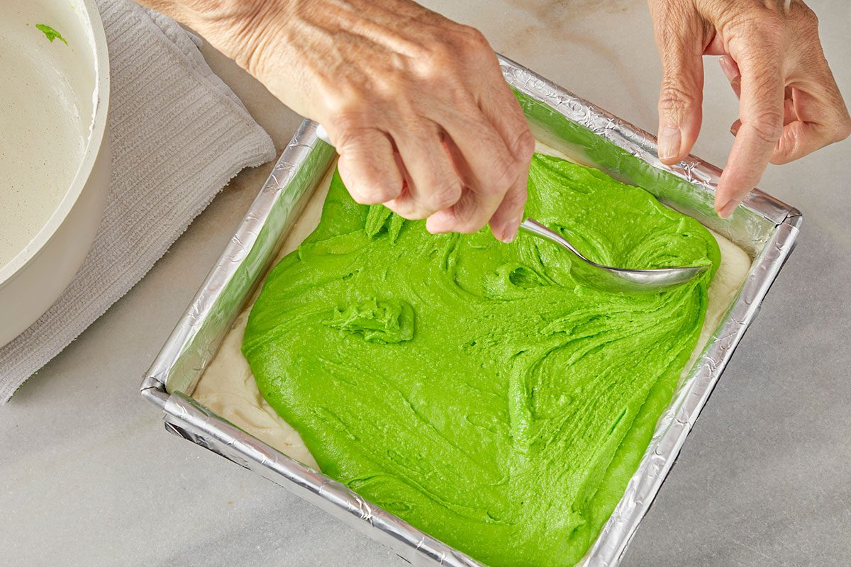 Overhead shot of a person uses a spoon to spread a layer of bright green batter over a white layer in a foil lined square baking pan; with a bowl and towel nearby on a marble surface