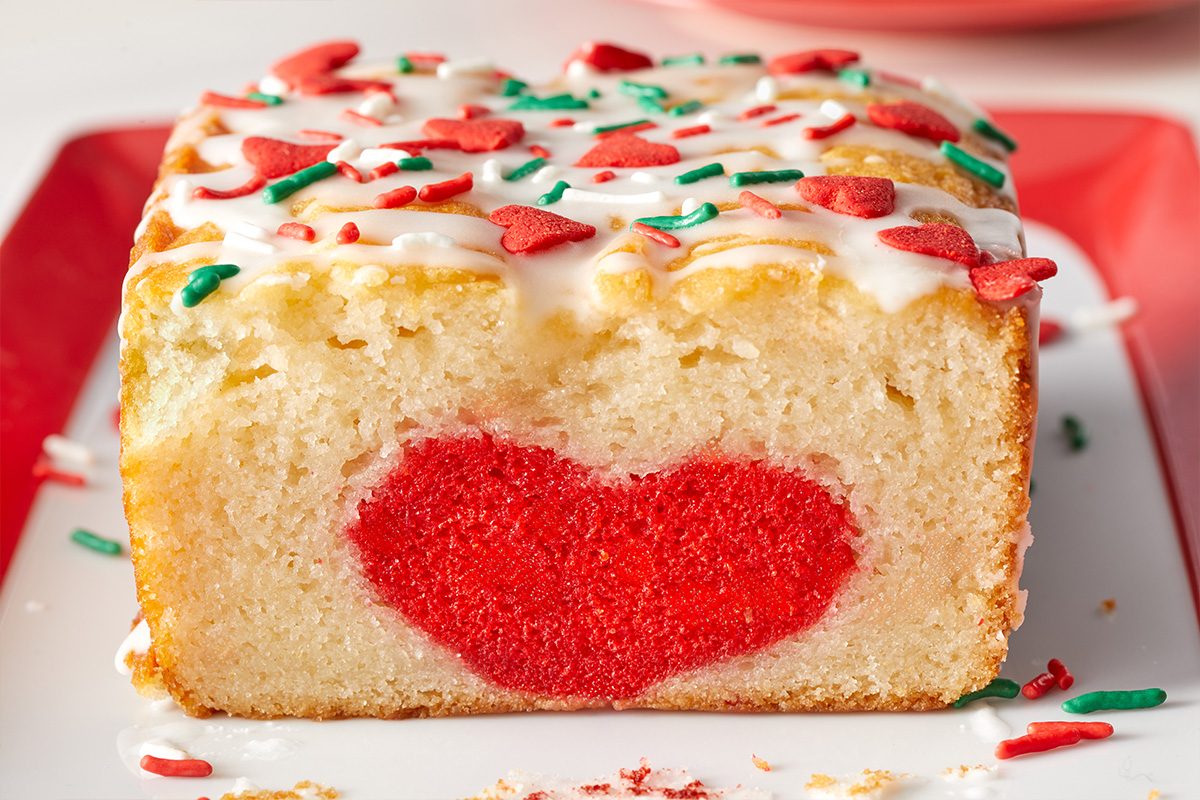 A loaf cake with white icing and red and green sprinkles sits on a plate. The cake has a red heart shape baked into the center, revealed in the sliced end facing the camera.