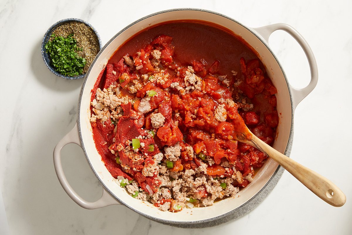 A large white pot filled with chili, containing ground meat, beans, tomatoes, and chopped vegetables, sits on a white surface. A wooden spoon rests in the pot, and a small bowl of chopped herbs and spices is nearby.