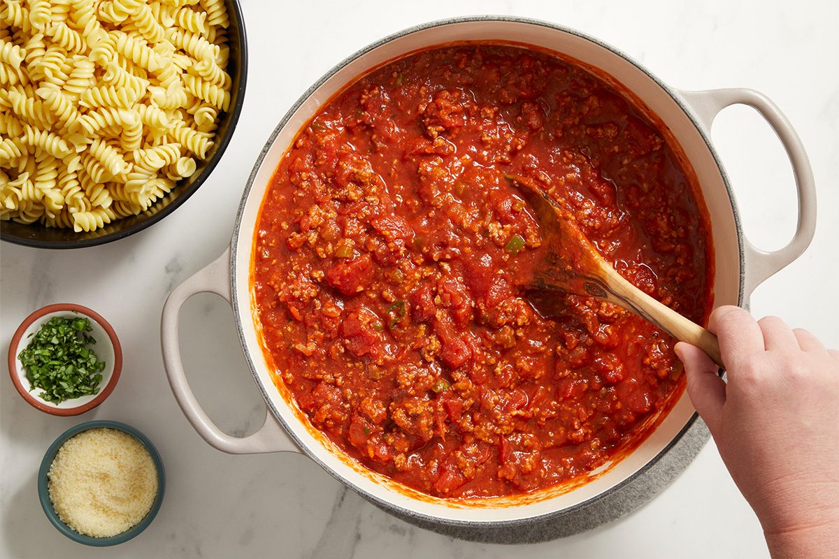 A hand stirs a pot of chunky red meat sauce with a wooden spoon. Next to the pot are bowls of uncooked rotini pasta, grated cheese, and chopped herbs on a white surface.