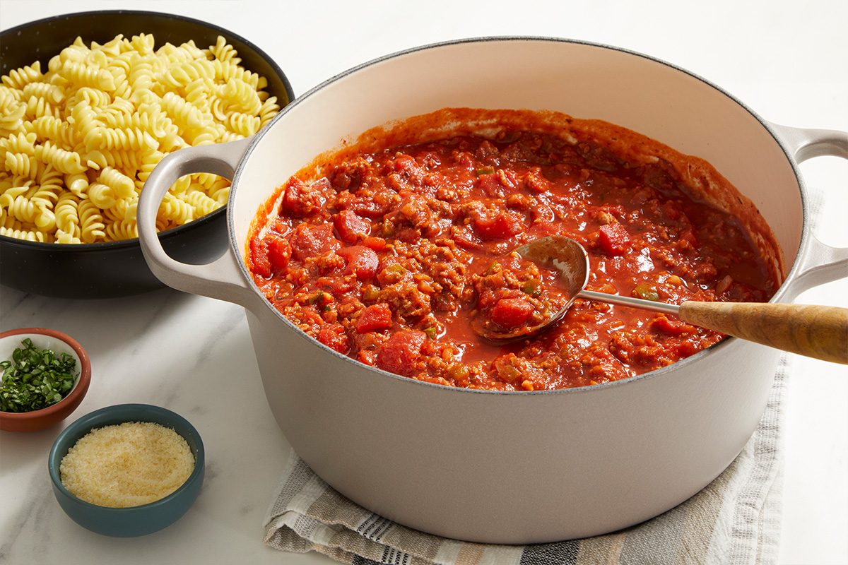 A pot of chunky tomato and meat sauce with a ladle inside sits on a cloth. Next to it are bowls of cooked rotini pasta, grated cheese, and chopped herbs on a white surface.