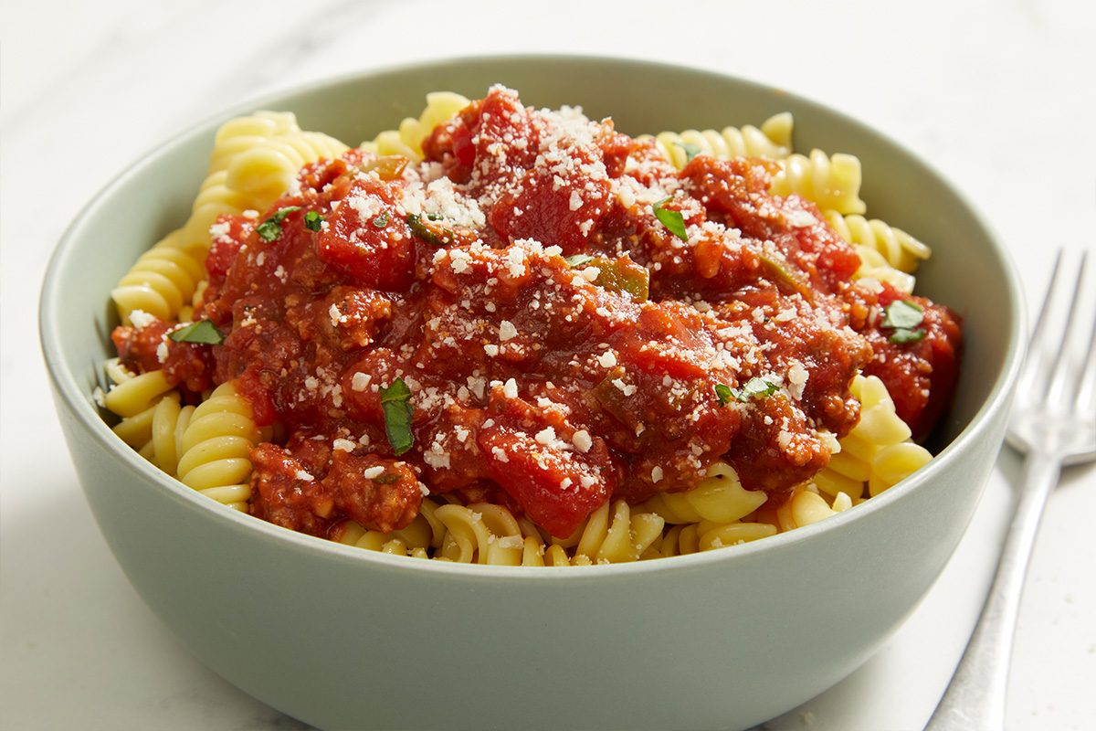 A bowl of rotini pasta topped with chunky tomato meat sauce and sprinkled with grated parmesan cheese, garnished with fresh herbs, sits next to a fork on a white surface.