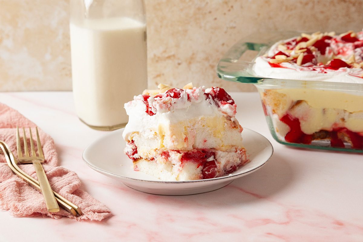 A slice of layered dessert with whipped cream and cherry topping sits on a white plate. Behind it are a glass baking dish with more dessert, a bottle of milk, and a pink napkin with two forks.