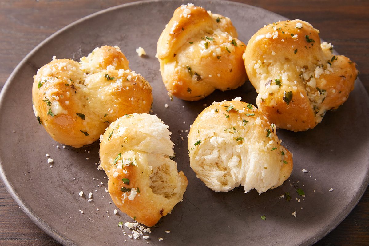 Four golden brown garlic knots sprinkled with herbs and grated cheese are arranged on a round gray plate, with one knot partially torn open to show its soft, fluffy interior.