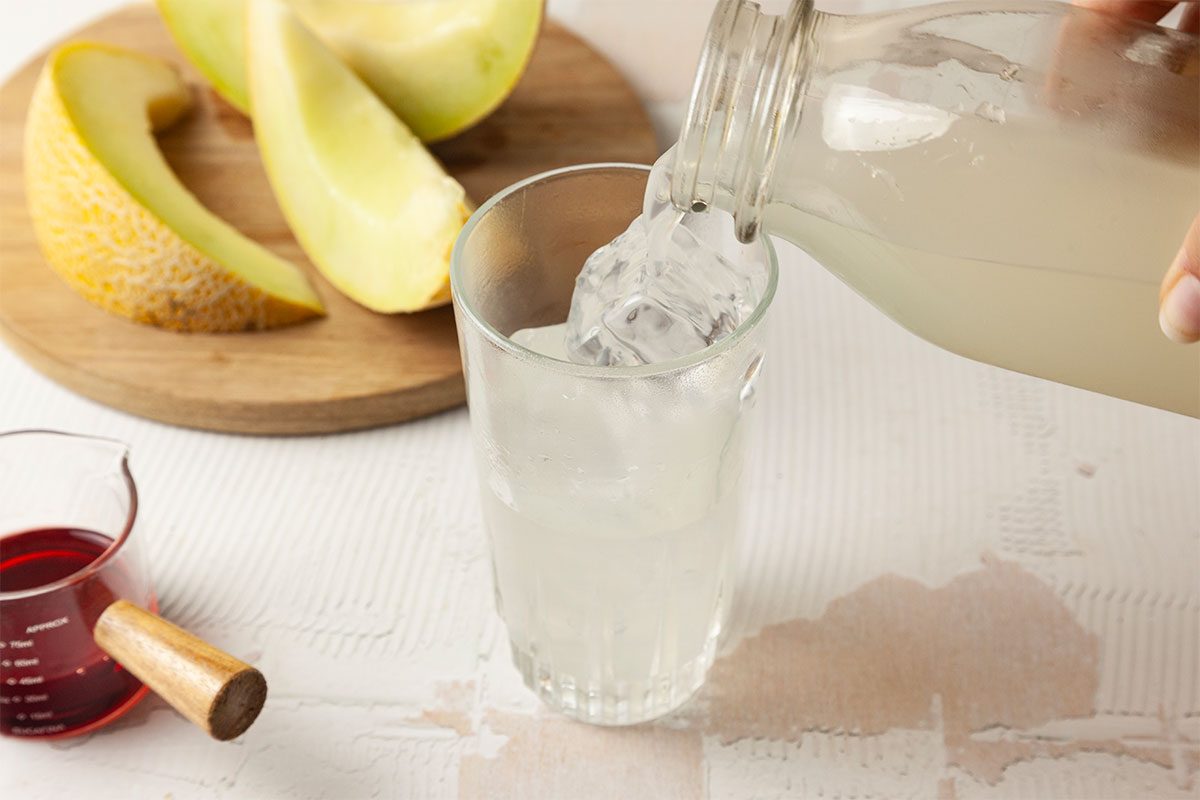 A hand pours a light-colored liquid from a glass bottle into a glass filled with ice. In the background, melon slices rest on a wooden board, and a small cup of red liquid sits nearby.