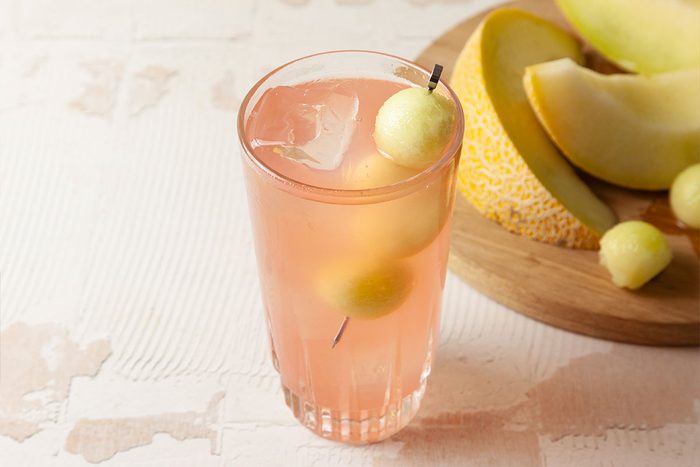 A glass of pink melon-flavored drink with ice and melon balls on a skewer, next to a wooden board with slices and balls of melon.