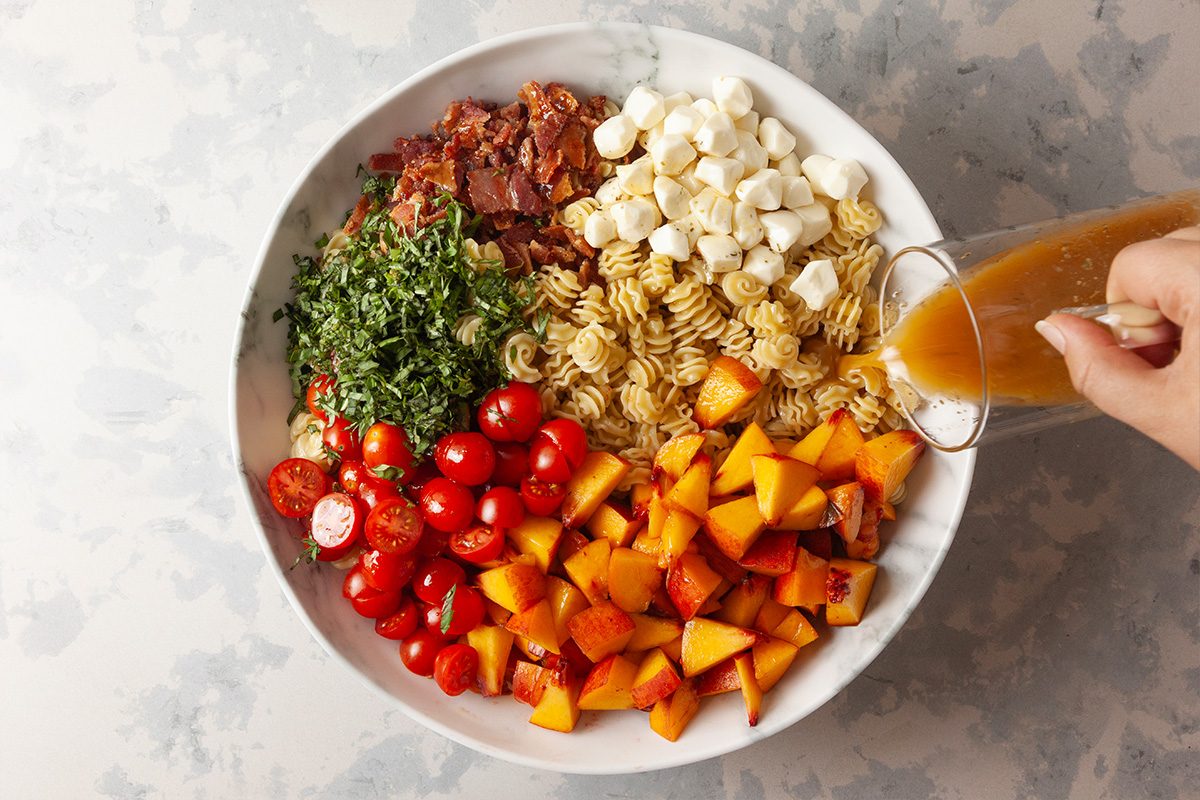 A large white bowl filled with pasta, chopped tomatoes, cubed peaches, mozzarella balls, chopped herbs, sun-dried tomatoes, and a hand pouring dressing over the ingredients.