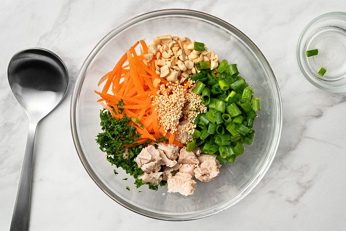 A glass bowl on a marble surface containing chopped green onions, shredded carrots, chopped peanuts, sesame seeds, cooked chicken pieces, and chopped herbs. A metal spoon and a small bowl are nearby.