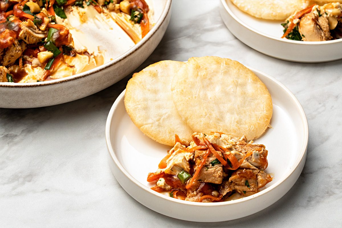 A white plate with two round, flat corn tortillas and a serving of shredded chicken in a tomato-based sauce with vegetables. Another plate and a serving dish are partially visible in the background.