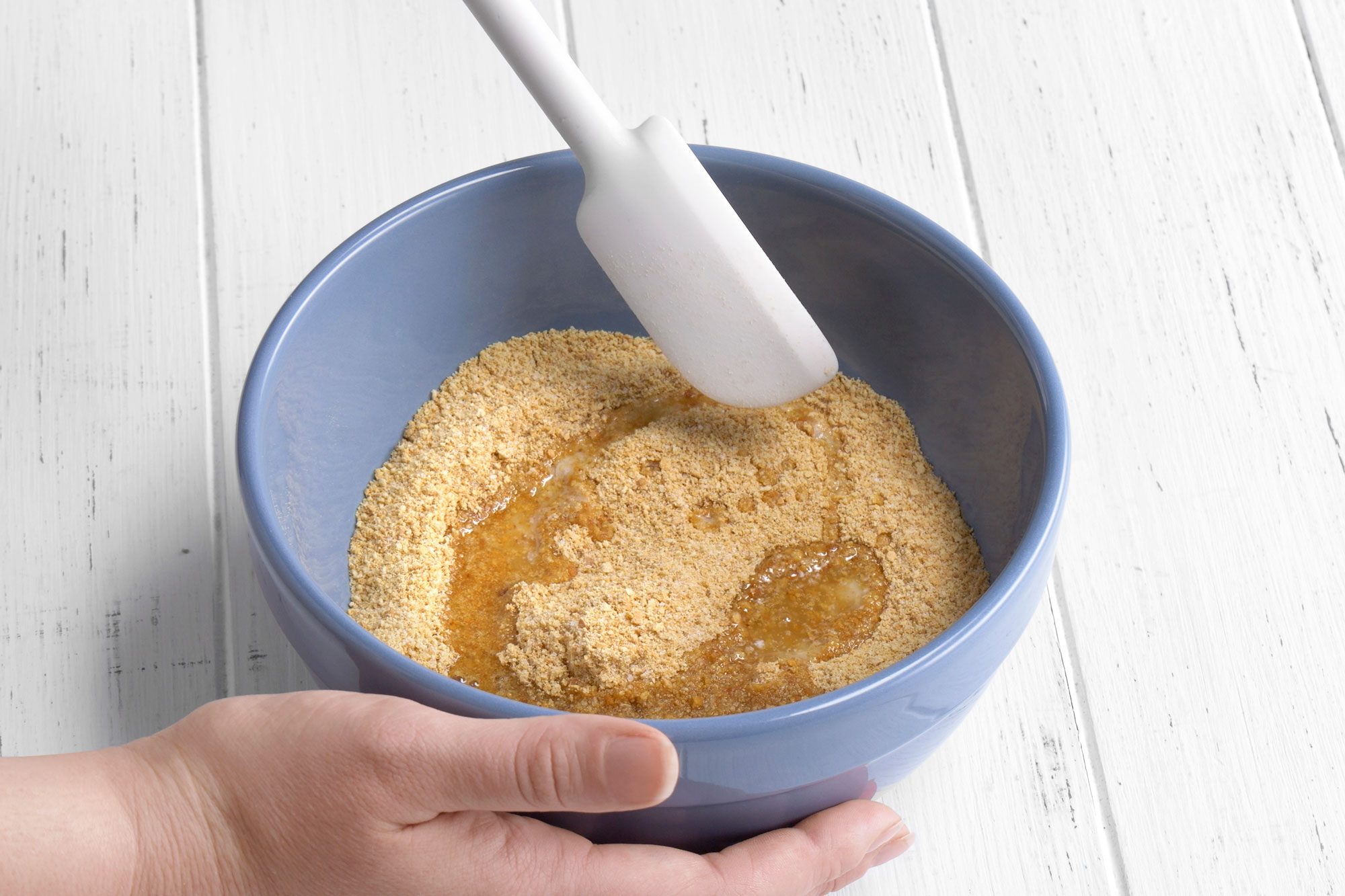 3/4th shot of a hand holds a blue bowl filled with crushed graham crackers and melted butter, while a white spatula is used to mix the ingredients on a white wooden surface
