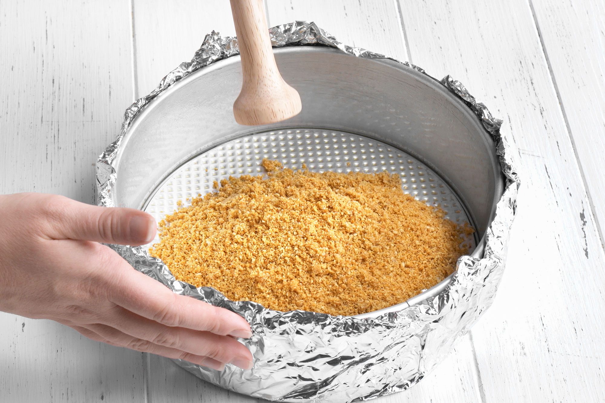 3/4th shot of a hand presses a layer of crushed crackers or biscuits into a foil wrapped springform pan using a wooden tamper, on a white wooden surface