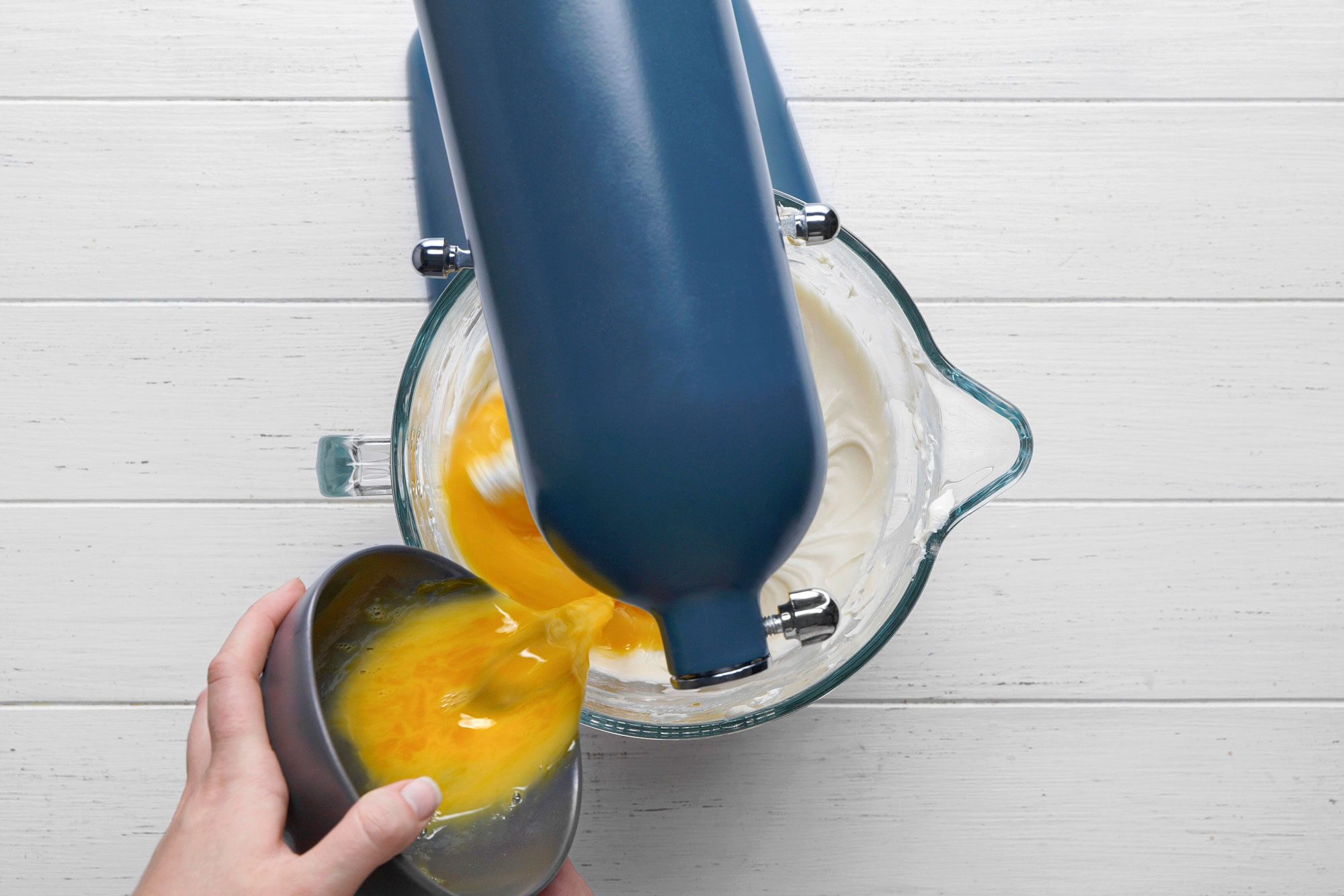 overhead shot of a hand pours beaten eggs from a bowl into a stand mixer with a glass bowl containing a white, fluffy mixture on a white wooden surface