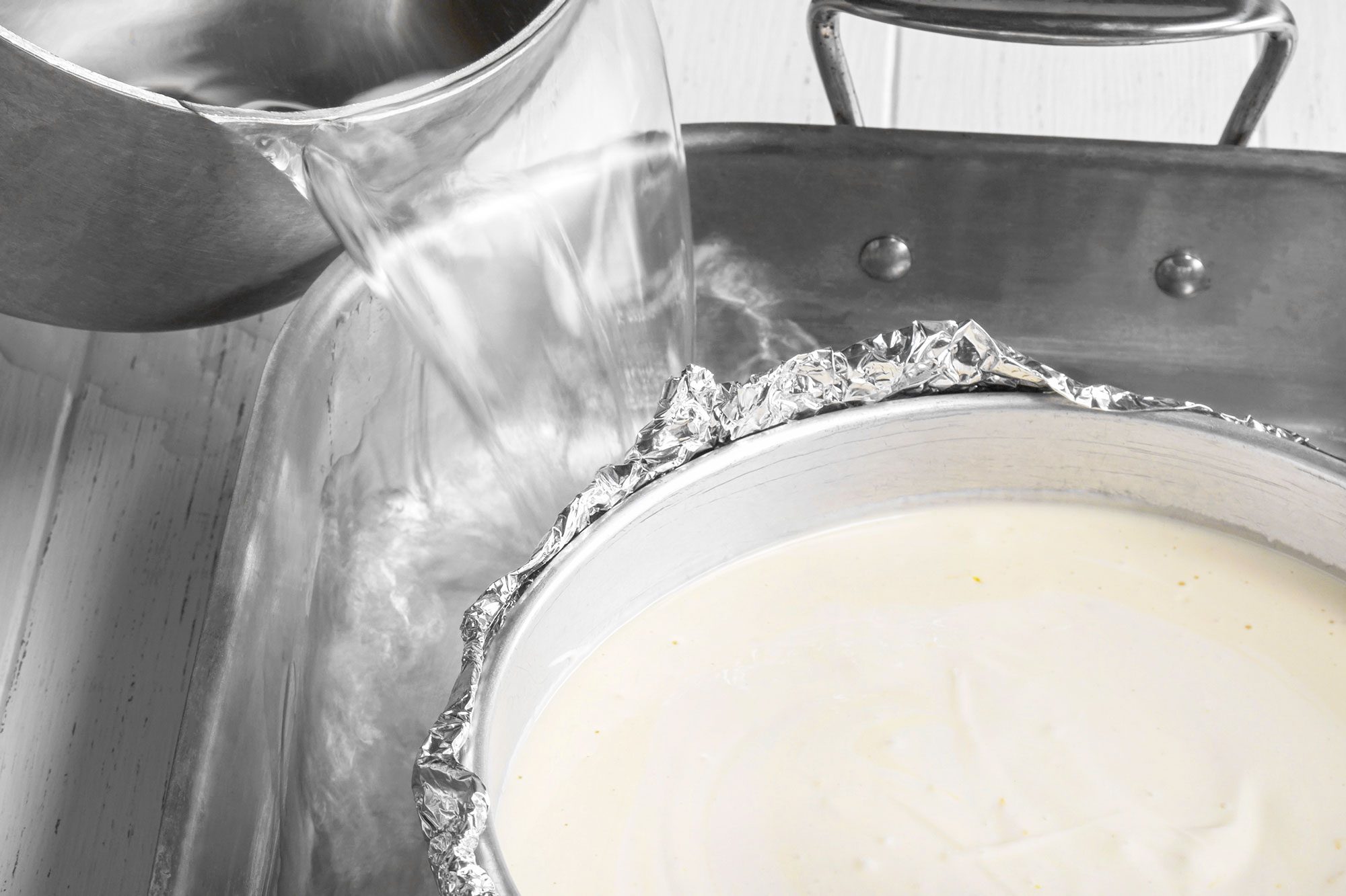 overhead shot of a person pours hot water from a saucepan into a large roasting pan lined with foil, surrounding a round cake pan filled with batter, preparing a water bath for baking