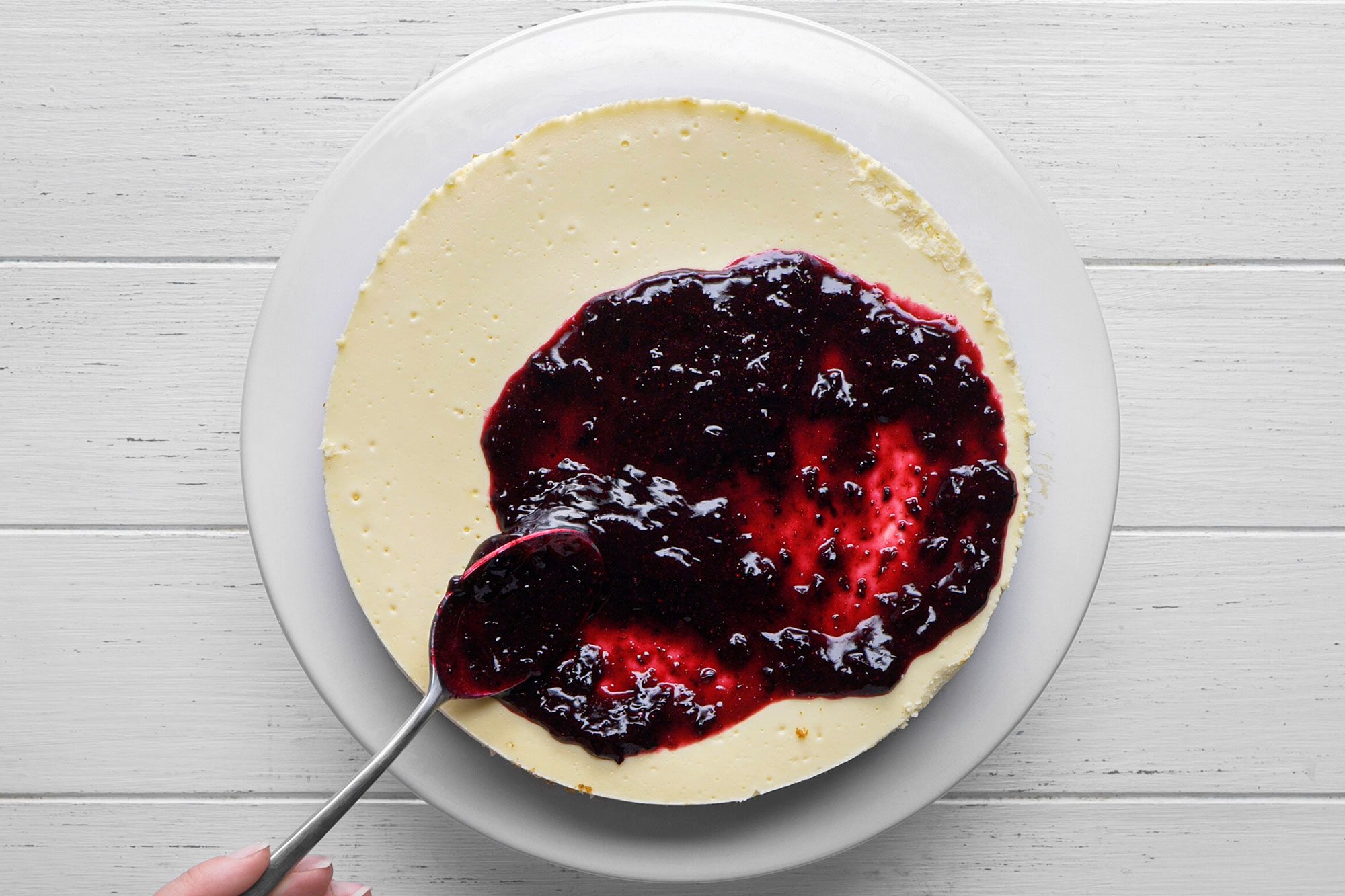 overhead shot of a hand is spreading a layer of dark berry sauce over a plain cheesecake on a white plate, set on a white wooden surface