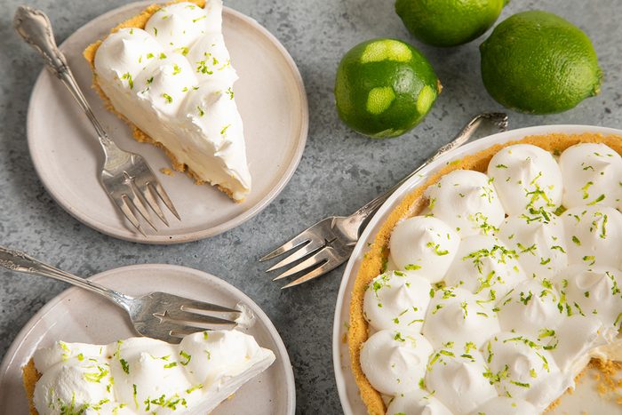 A key lime pie topped with whipped cream and lime zest is served on two plates with forks. Whole limes are nearby on a gray surface.