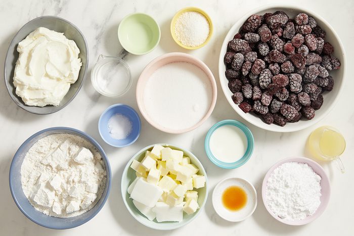 An assortment of baking ingredients in bowls on a white surface, including frozen berries, flour, sugar, butter cubes, cream cheese, milk, vanilla extract, lemon juice, and small bowls of salt and baking powder.
