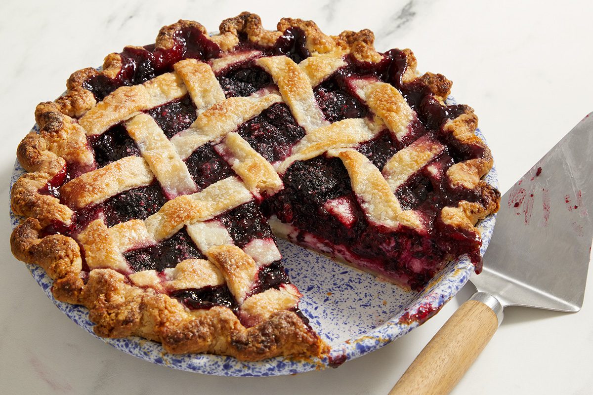 A freshly baked berry pie with a golden brown lattice crust sits in a pie dish. A large slice has been removed, and a pie server with a wooden handle rests beside it.