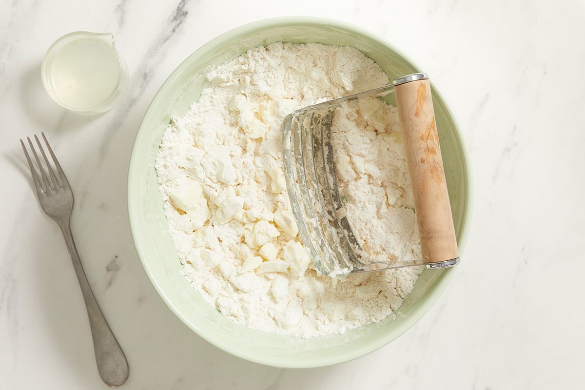 A bowl of flour being mixed with a pastry cutter on a white surface, with a fork and a small cup of liquid nearby.