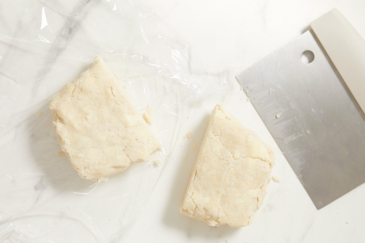 Two pieces of dough sit on a sheet of plastic wrap next to a metal bench scraper on a white marble surface.
