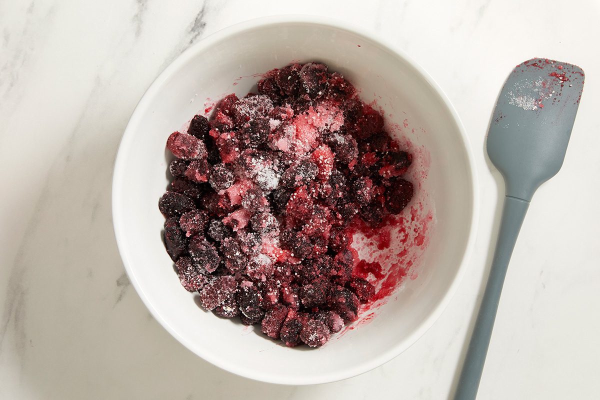 A white bowl filled with frozen berries coated in sugar sits on a white marble surface next to a gray spatula with berry stains.
