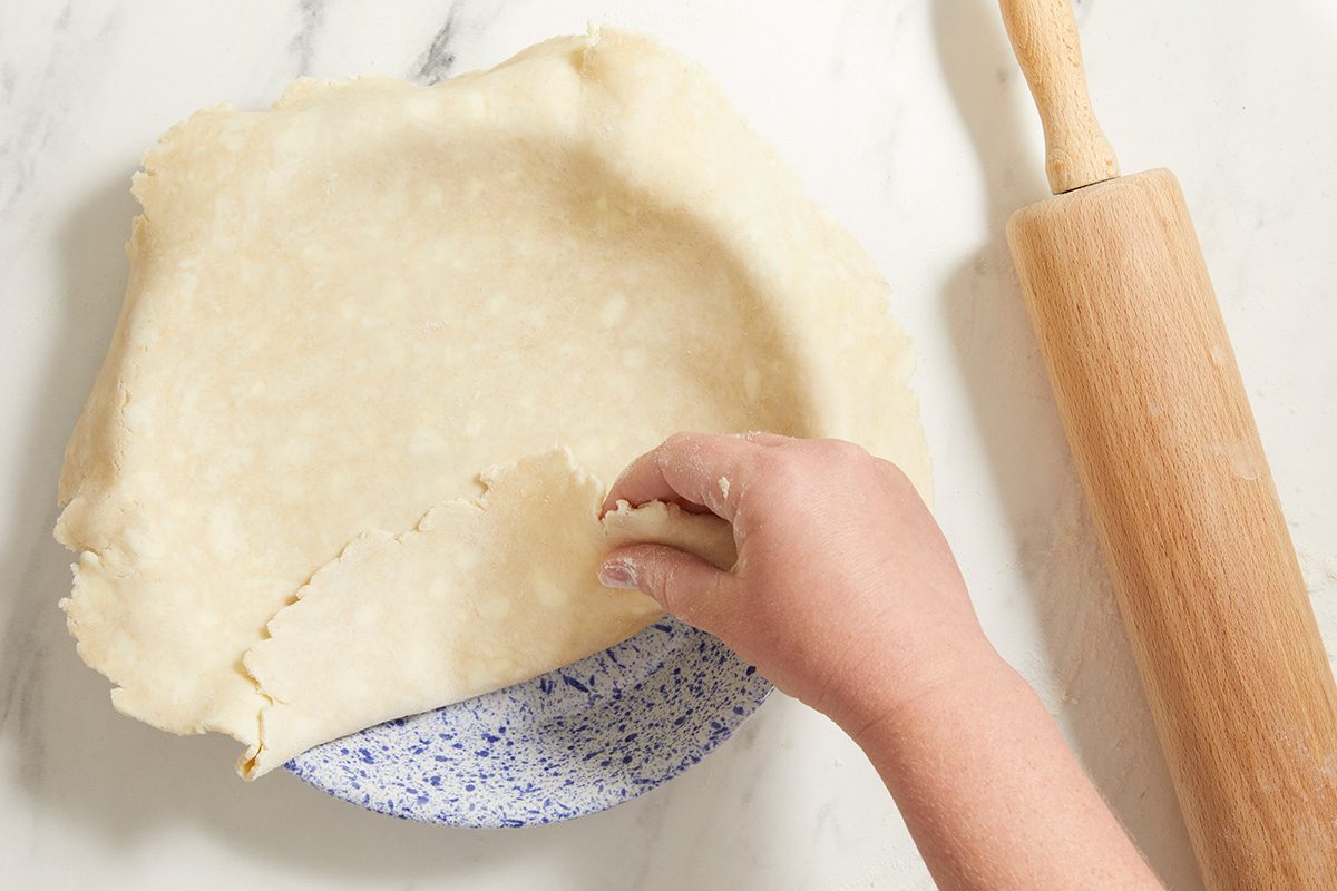 A hand lifts the edge of raw pie dough draped over a speckled pie dish on a marble surface, with a wooden rolling pin lying beside it.