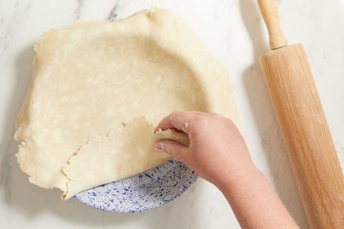 A hand lifts the edge of raw pie dough draped over a speckled pie dish on a marble surface, with a wooden rolling pin lying beside it.
