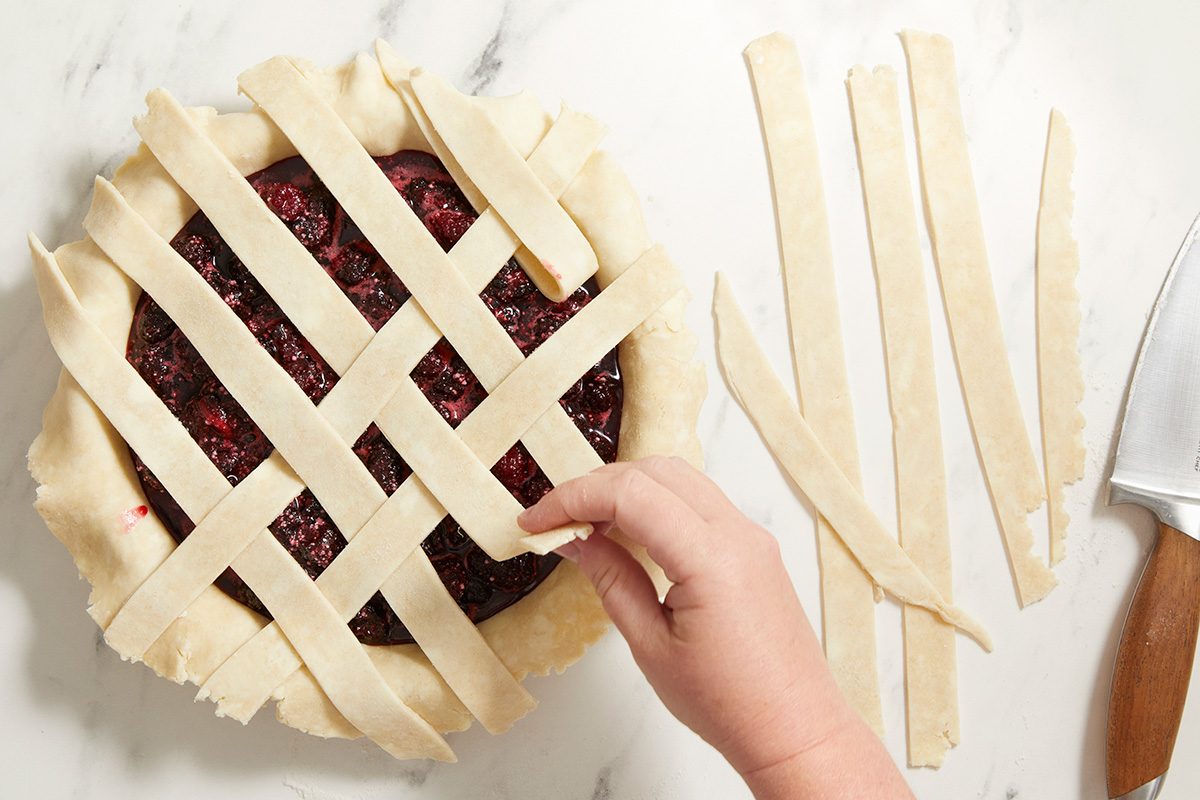 A hand is weaving strips of pie dough into a lattice pattern over a fruit pie filling on a marble surface, with extra dough strips and a knife nearby.
