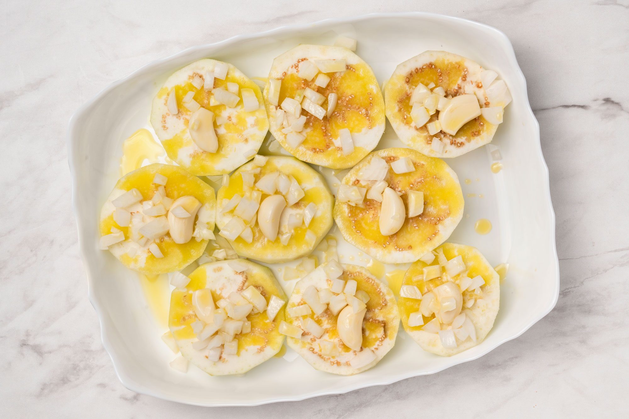 Overheda shot of a white dish with sliced eggplant topped with chopped onions, whole garlic cloves, and drizzled olive oil, set on a light marble surface;